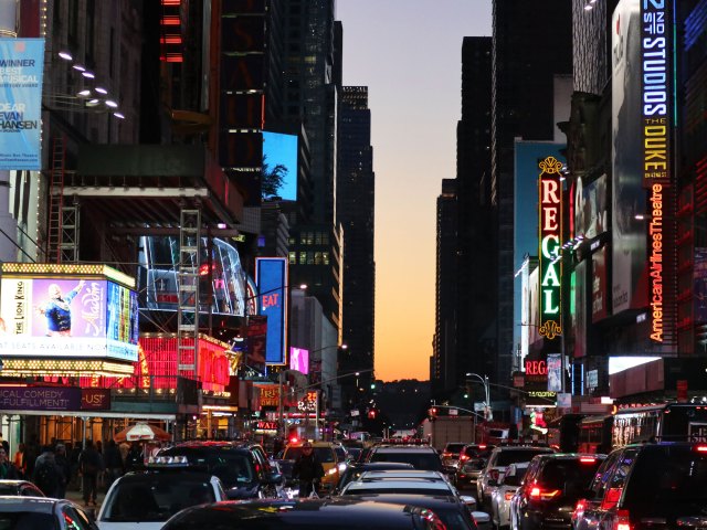 Traffic-filled New York City street lined with neon billboards and theater marquees at sunset