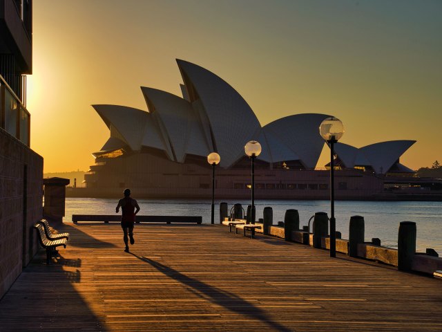 The silhouette of a man running on a dock at sunset with the white sails of the Sydney Opera House in the background