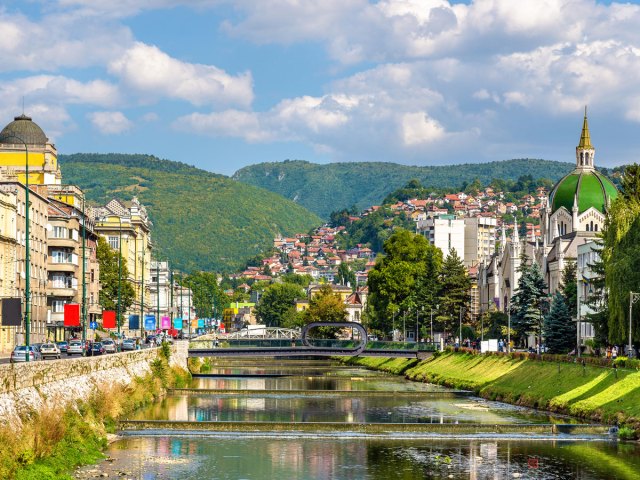River flanked by buildings with mountains in background in Bosnia and Herzegovina