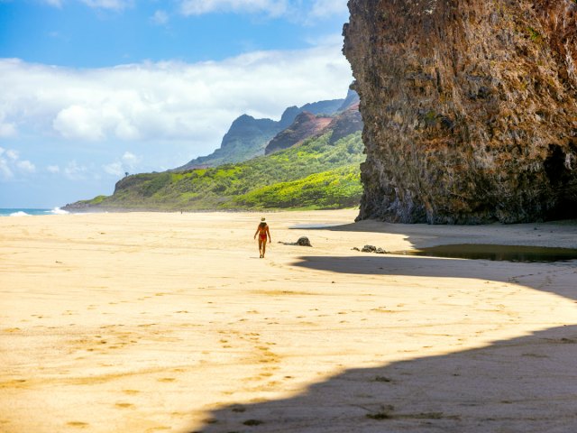 Person walking in the shadow of tall sea cliff on sandy beach on Kauai