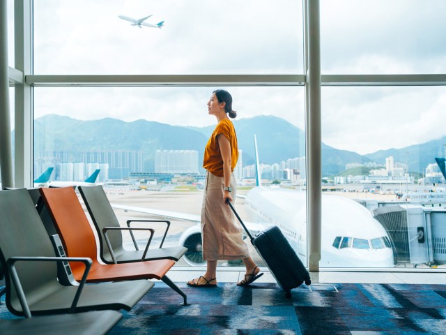 Woman rolling suitcase through airport, with view of airplanes through window