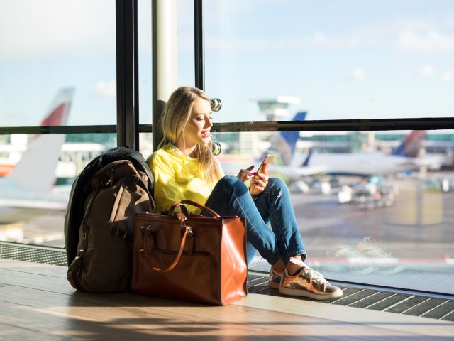 Traveler browsing phone while sitting on airport floor next to windows overlooking tarmac