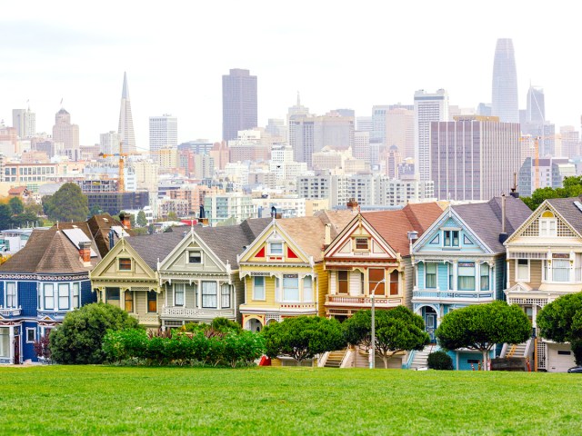 Colorful Victorian row homes seen across park, with San Francisco skyline in the distance