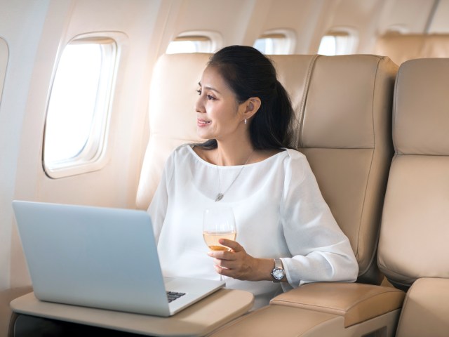 First class passenger looking out airplane window holding glass of wine with laptop open in front of her