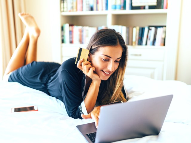 Person laying on bed using laptop and holding credit card in hand