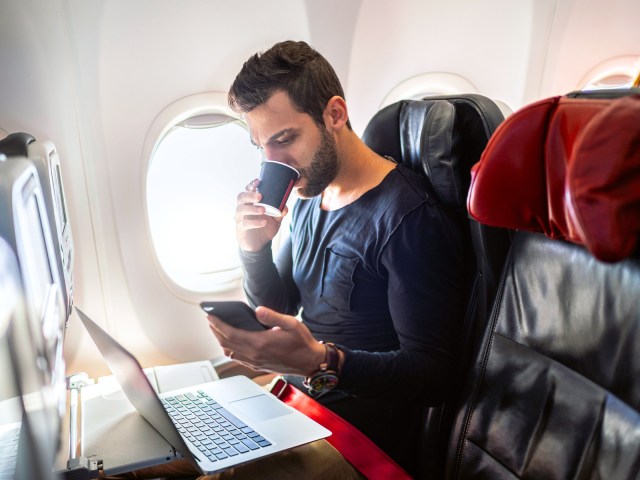 Man in airplane seat drinking coffee and working on laptop on tray table