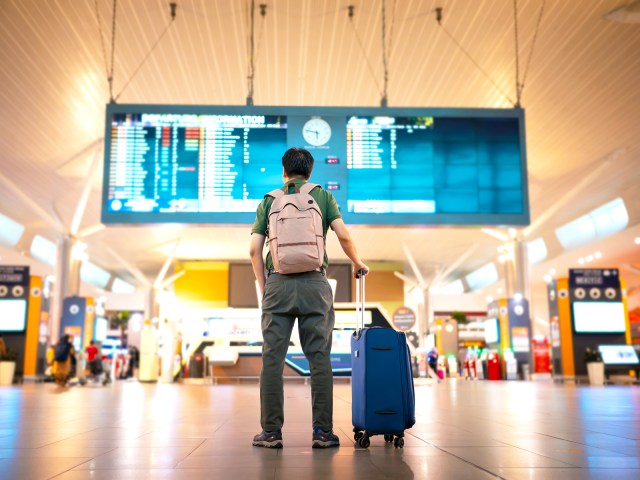 Traveler with backpack and suitcase looking at departures board in airport