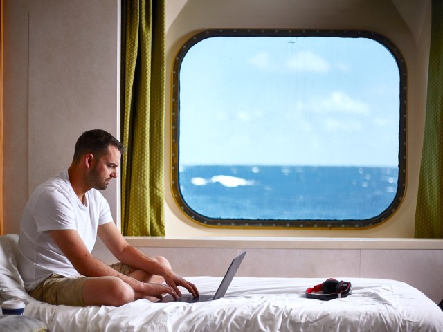 Cruise ship passenger working on laptop in bed, with view of sea out cabin window