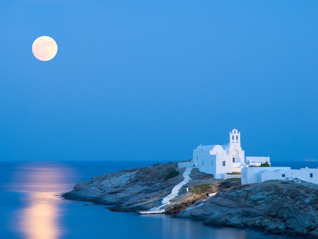 White church on rocky peninsula with moon visible on Sifnos, Greece